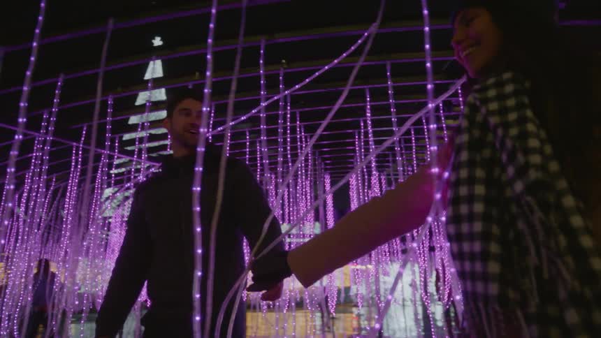 Man and woman walk through purple string christmas lights at night