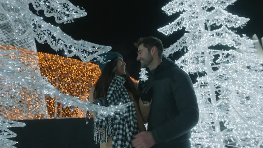 A woman touching a christmas tree while a man runs up and hugs her