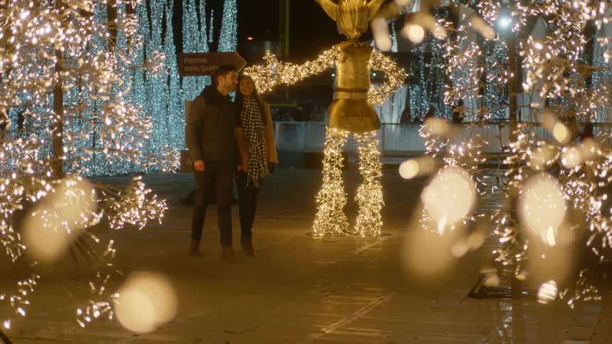 A man and woman hold hands while walking through an outdoor plaza filled with sculptures made out of christmas lights