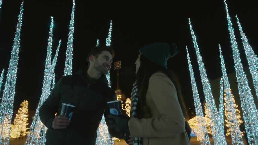 A man and woman hold their coffees as they walk near some christmas lights