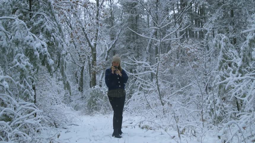 Woman stands in the forest and uses smartphone while it snows during daylight