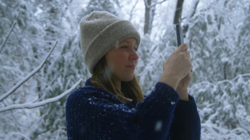 A young adult female smiles while taking a photo with her phone outside in the snow