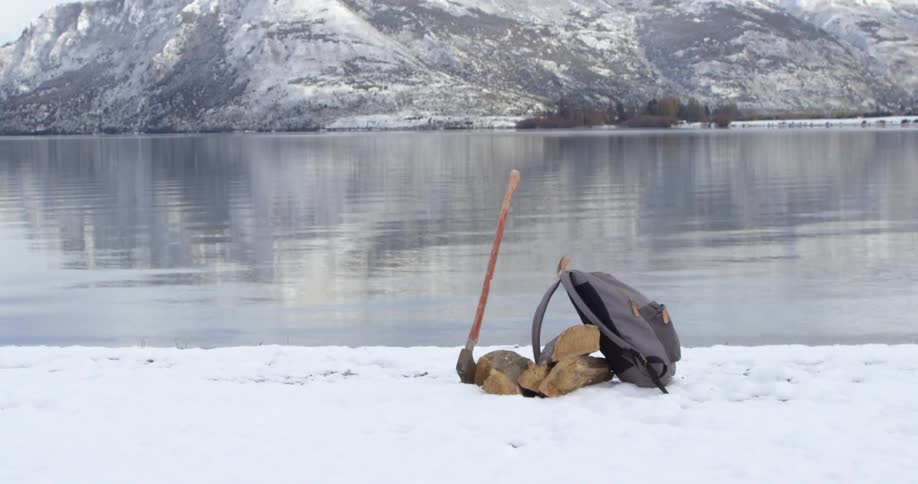 A backpack and axe stand against some rocks in the snow near a body of water