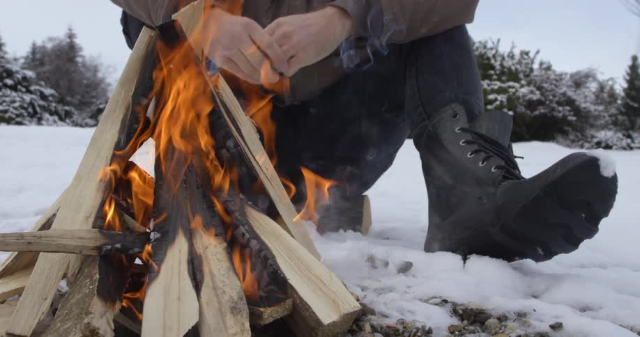 A man sits on a small log, warming his hands next to a campfire on an overcast winter day