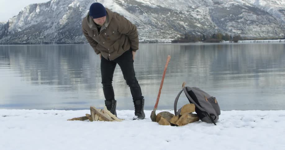 A white man carries kindling to the shore of a lake in the winter