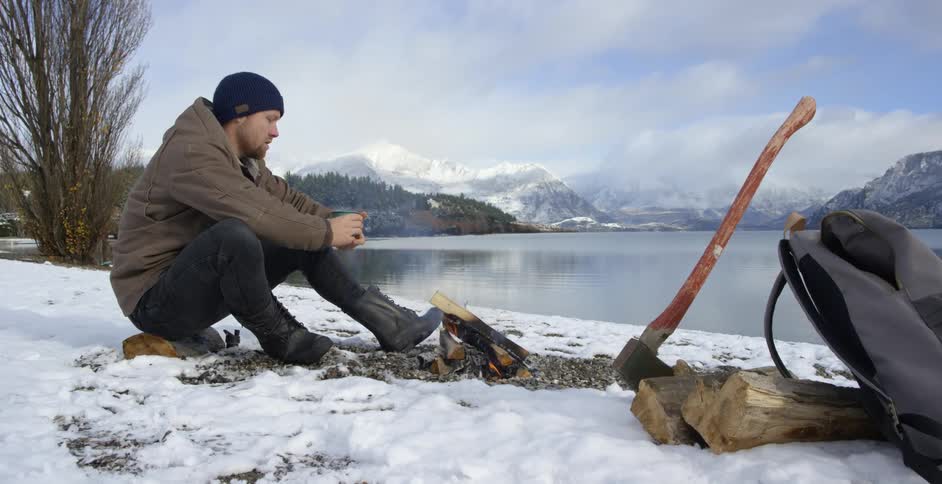 Man sips warm beverage by campfire surrounded by snow