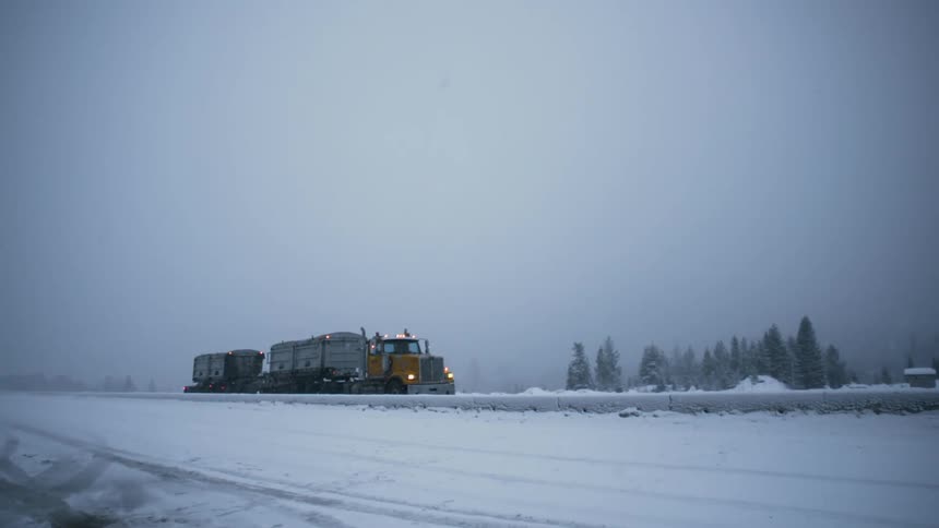 A semi-truck drives through a rural area during a snowstorm