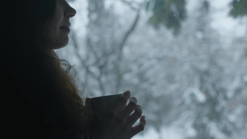 A woman holds a mug as she looks out at the trees and snow