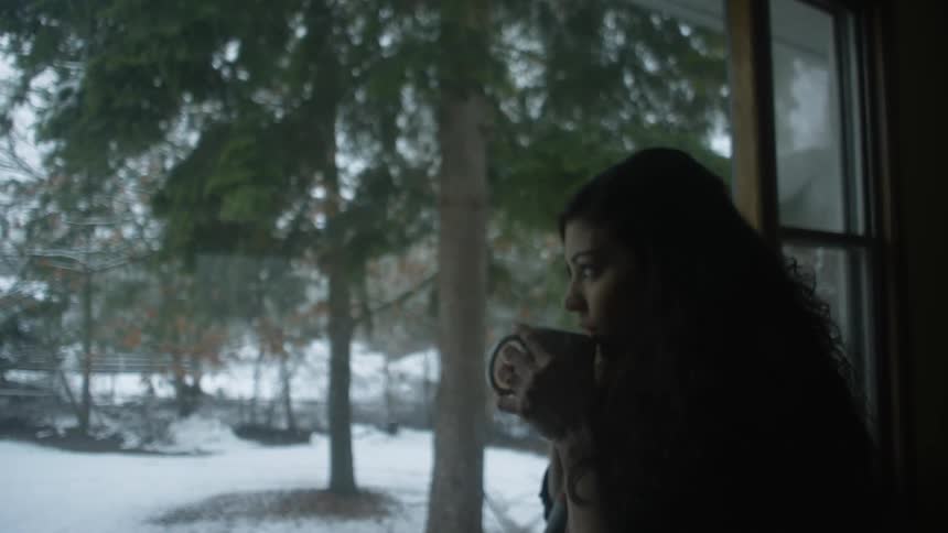 A woman sips from a mug while looking out of a window on a cold winter day