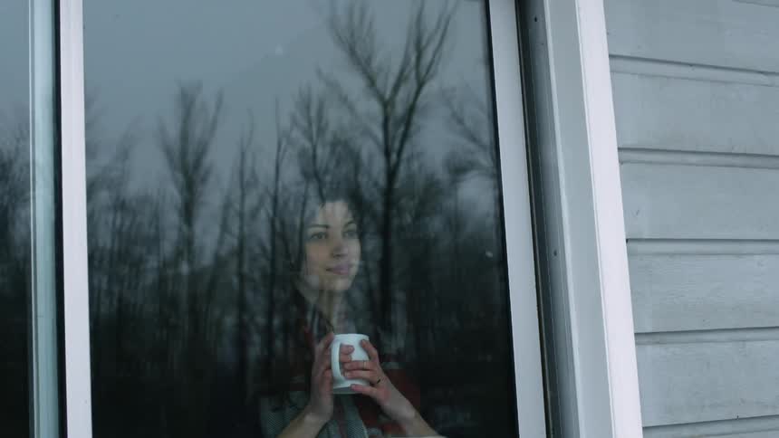 A woman holding a coffee mug stands looking out a house window at falling snow