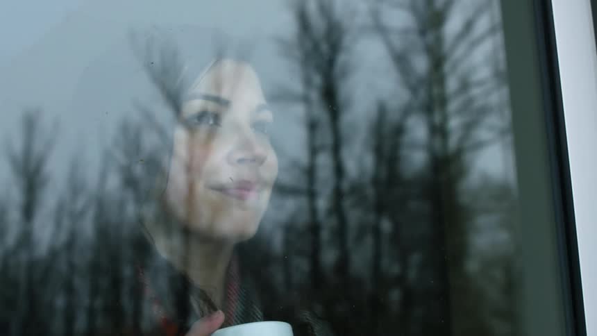 A woman looks out of a window which reflects bare trees