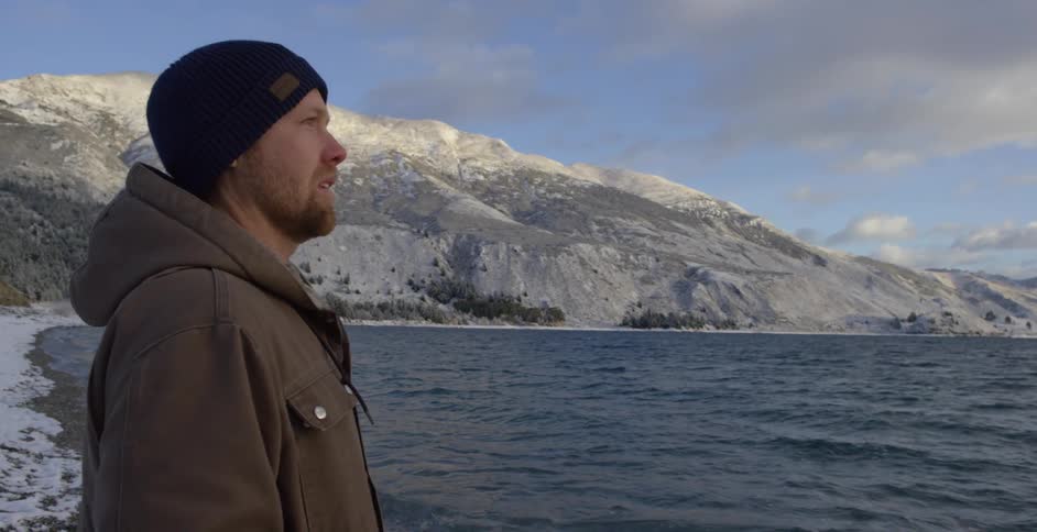 Man standing lakeside on a snow-covered bank looking out over the water