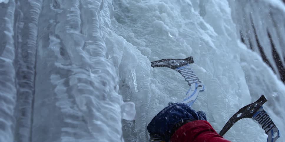 A climber is using ice axes to scale a wall of ice during the day