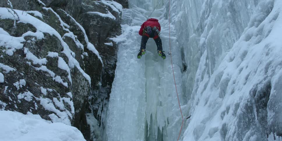 The snow is falling as this person climbs the ice on the cliff