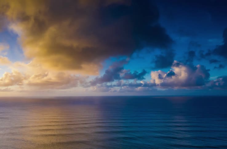 Fluffy clouds are passing over ocean water as the sun sets