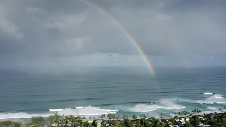Aerial view of rainbow and ocean waves breaking against the beach of coastal town on cloudy day