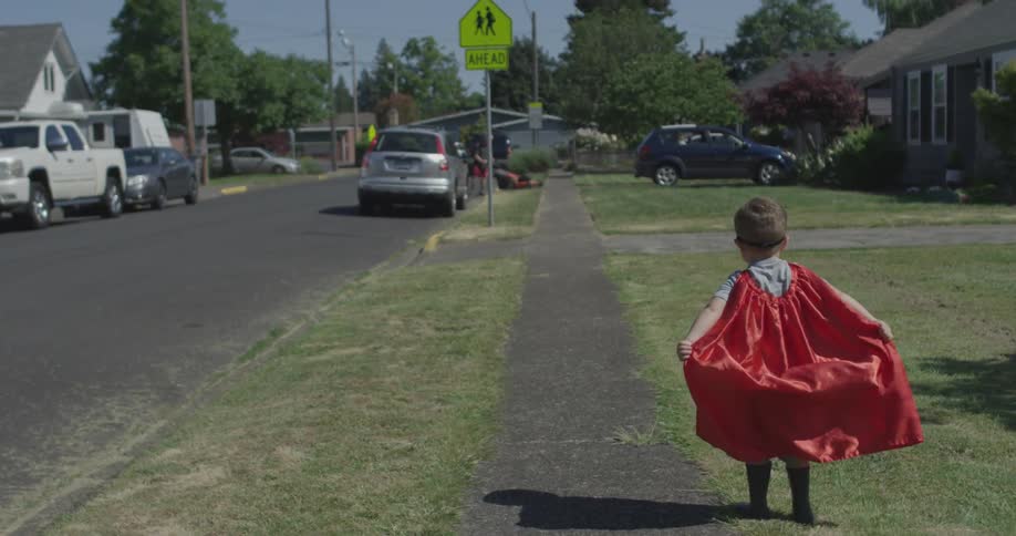 A boy raises his red cape as he stands by a sidewalk