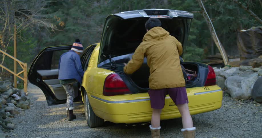 A man quickly puts suitcases in the trunk of a taxi while his wife and two children get in the backseat and then he runs and gets in the front seat