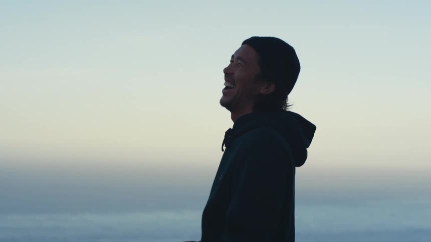 A man is standing on a hill overlooking the ocean and holding a mug and laughing