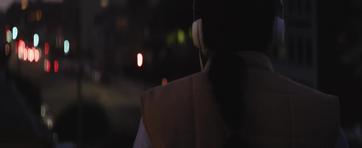 A woman wearing headphones outside stops to look at passing traffic at night