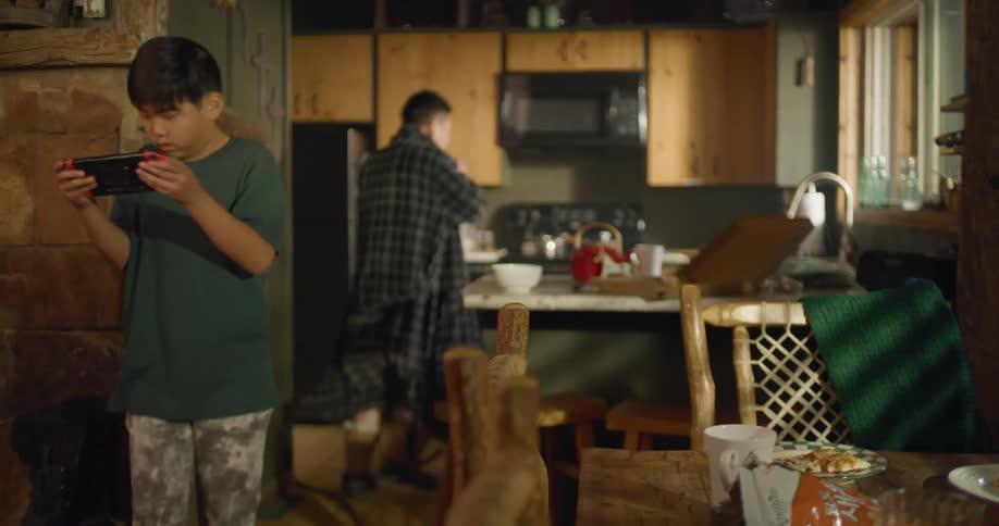 A man in his home is rushing around while brushing his teeth and his son is casually walking around staring into a smartphone in the daytime