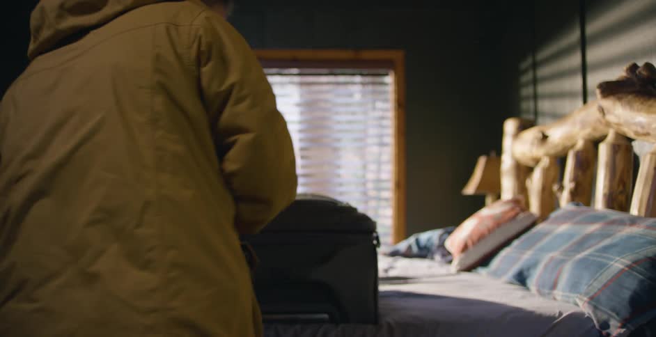 A boy wearing a raincoat is standing over a bed as he closes and zips a suitcase and picks it up and leaves