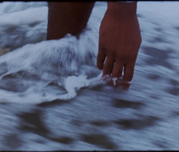 Someone is on a beach and drops a yellow rose into the water as the waves roll in