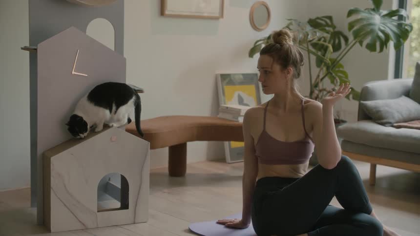 Young woman on a yoga pose over a mat in the living room looking at her cat