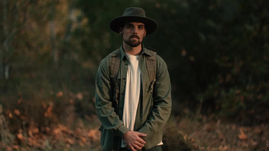 Young bearded man with a hat standing outdoors at sunset looks at the camera