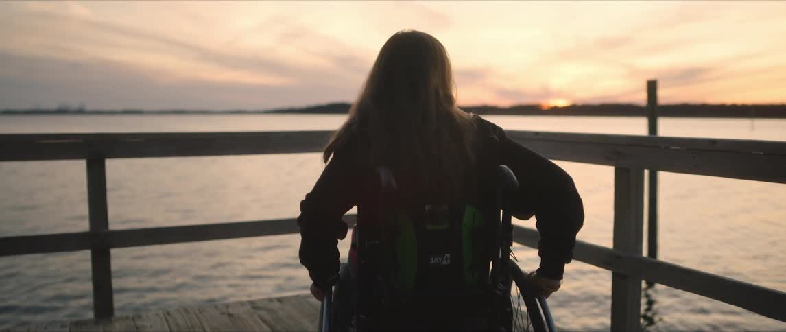Woman in wheelchair maneuvers toward wooden rail with view of red sky over opposite shore