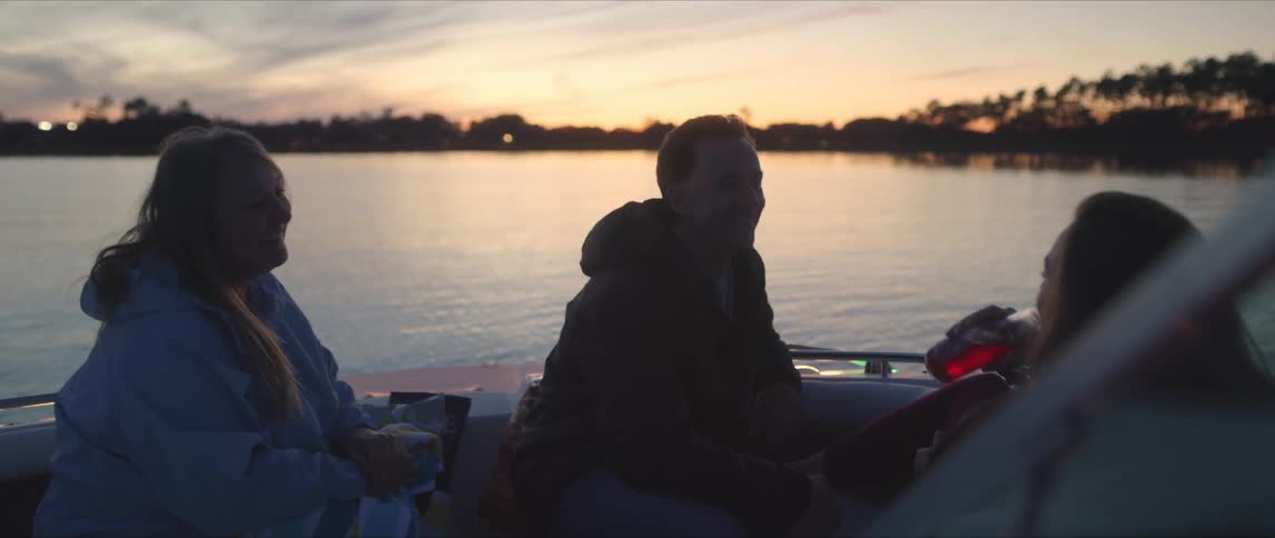 Three friends sitting on a boat laughing and drinking as the boat floats near shore at sunset