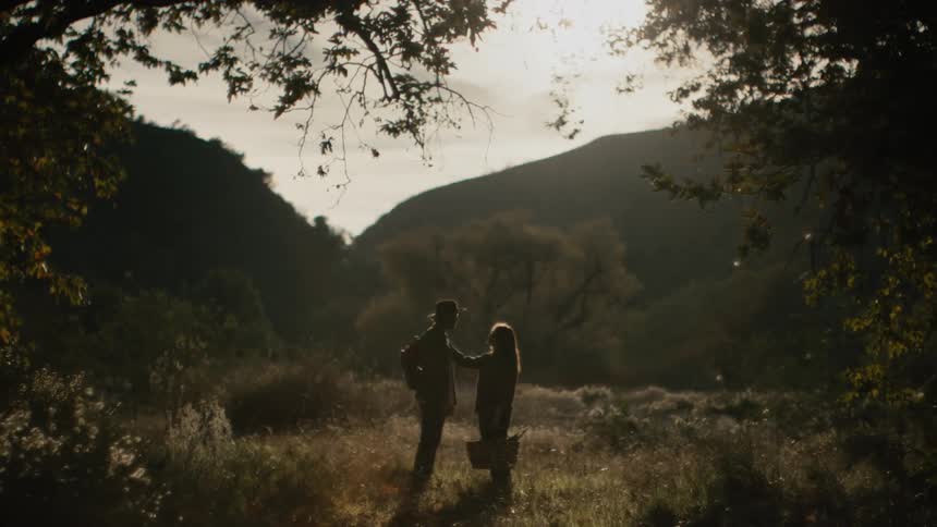 A man wearing a hat and a backpack is talking to a woman holding a picnic basket and they reach out to each other and kiss and there are mountains in the distance