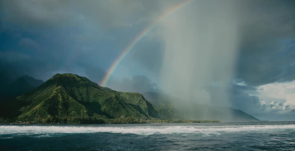 Waves roll in as a rainbow stretches across the sky over a mountain