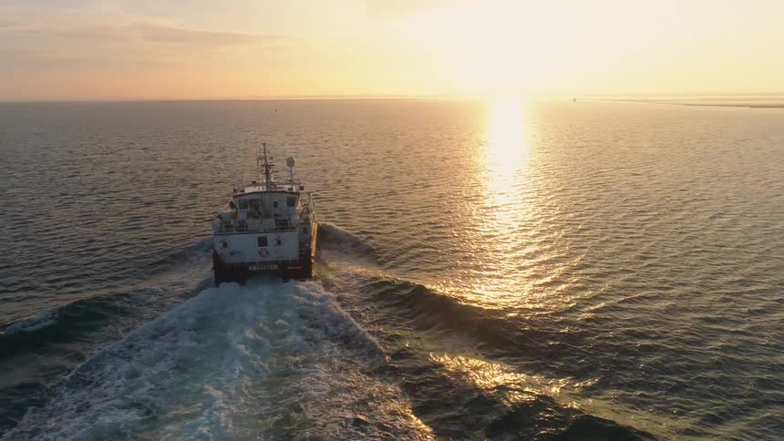 Large boat plows through water leaving wake as it heads toward horizon as ocean surface reflects sun sitting on horizon