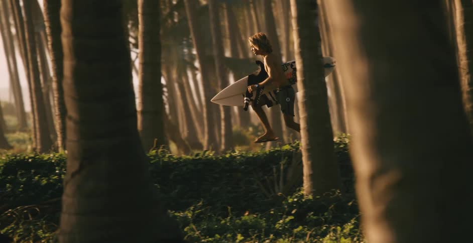 A young man wearing shorts is carrying a surfboard and running through a grove of palm trees