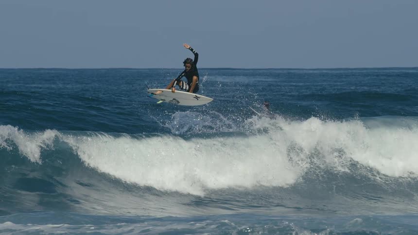 A surfer riding a curling wave jumps and turns in midair while others paddle out to catch a wave