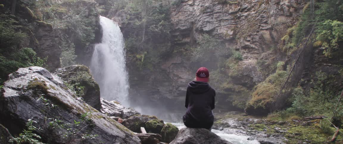 Woman with red cap and black coat sits on rock watching waterfall cascading down cliffside