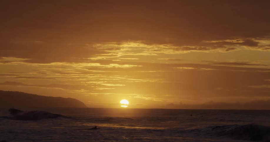 Waves roll in under an orange sky as the sun sets on the horizon