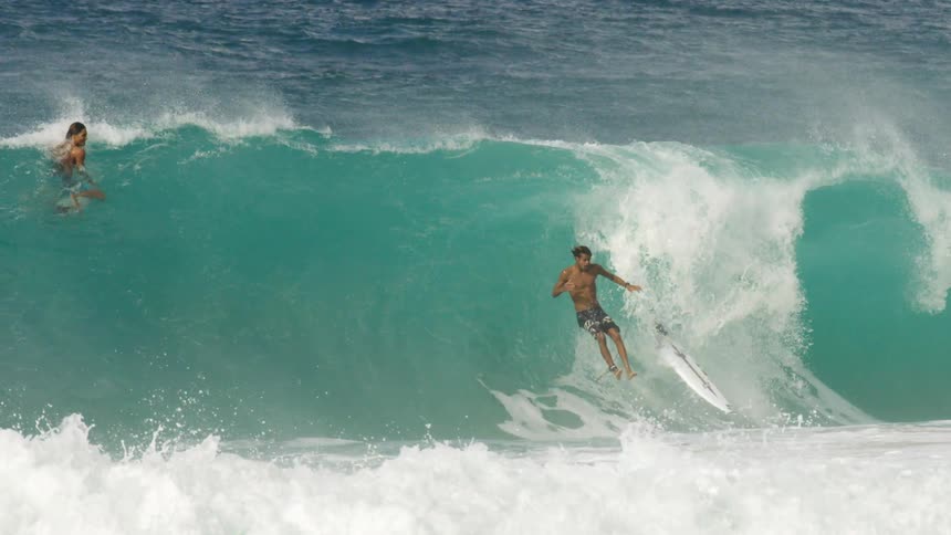 Three men surf in the ocean as one man tries to stand up on a large wave and falls off his board