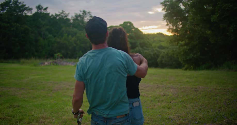 Woman with black blouse and jeans kisses man with dark blue cap turquoise t shirt jeans holding each other and making out in the countryside at dusk