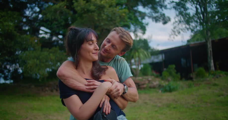 Man with turquoise t shirt holds woman with brown wavy hair and black dress lovingly looking into each other and making out in the countryside at dusk