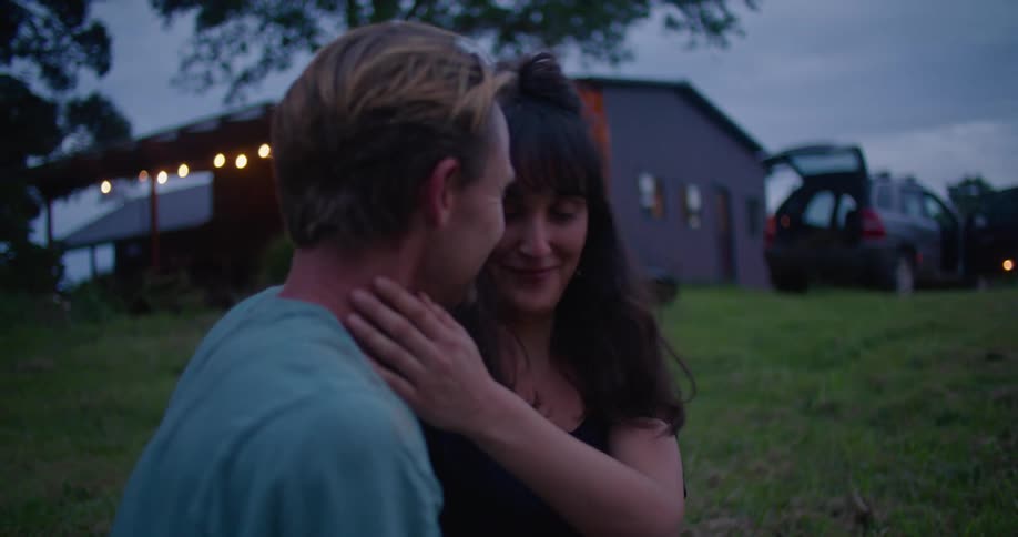 Man with turquoise t shirt sitting on green grass with woman with brown hair and black blouse holding and kissing each other in the countryside at dusk