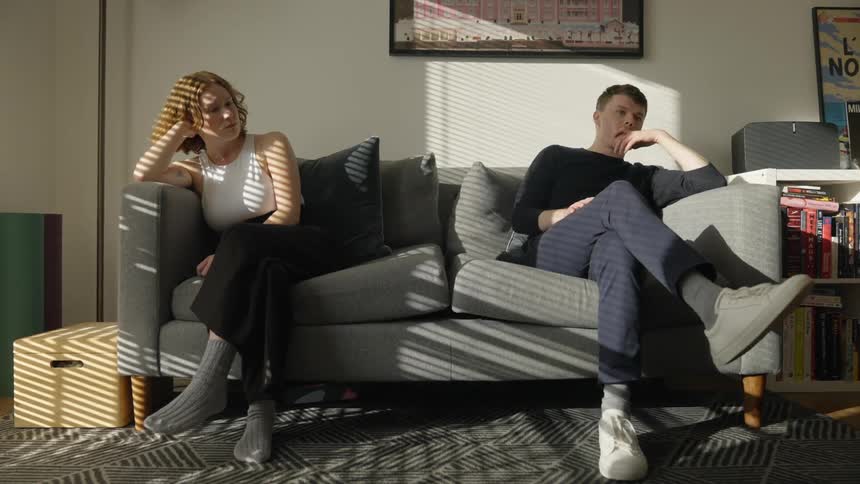 Young man sits besides a woman on a gray couch at home not looking at each other