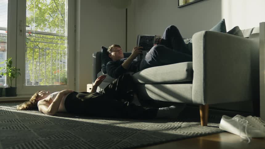 A woman who is laying on the floor reaches up to a man laying on a couch reading a book