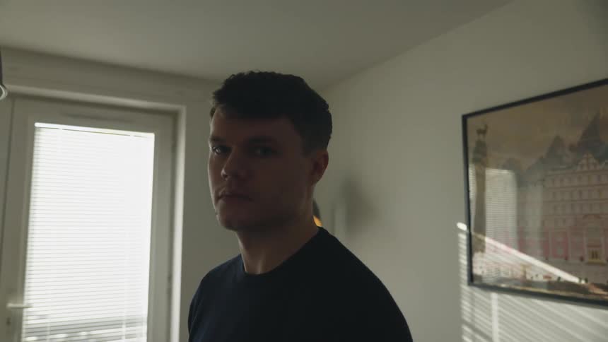 A man standing in the living room of a house turns to look solemnly into the camera