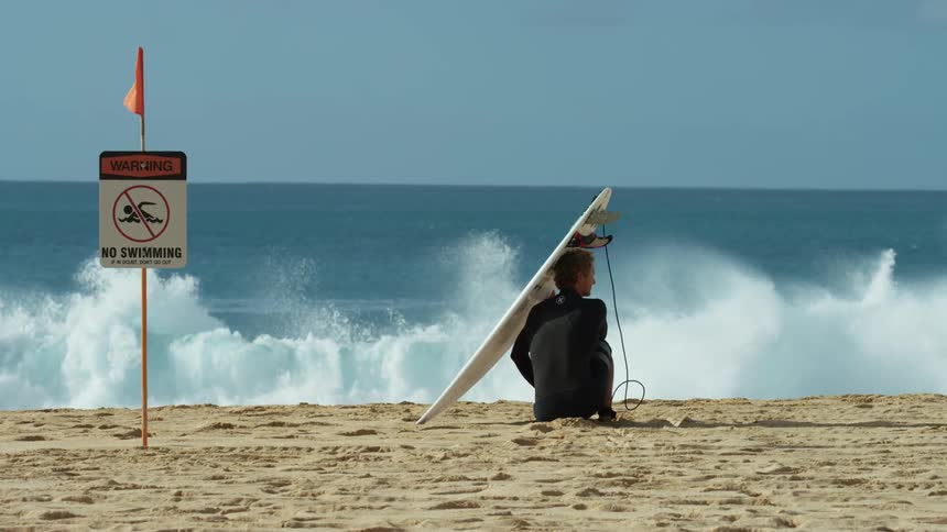 Surfer sitting on the shoreline watching waves crash next to a no swimming sign