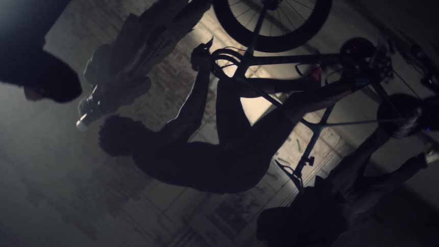 Silhouette of a man riding a static bicycle surrounded by photographers inside an abandoned warehouse