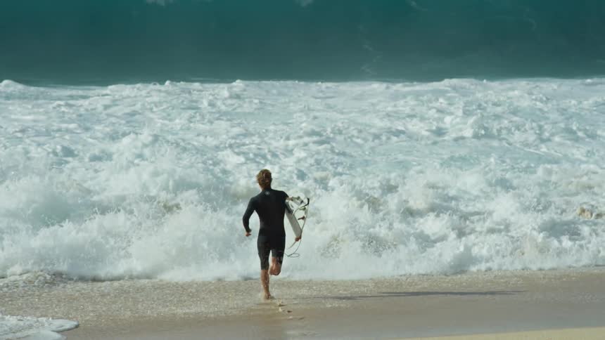 A man runs into the ocean with his surfboard and jumps on to catch a wave