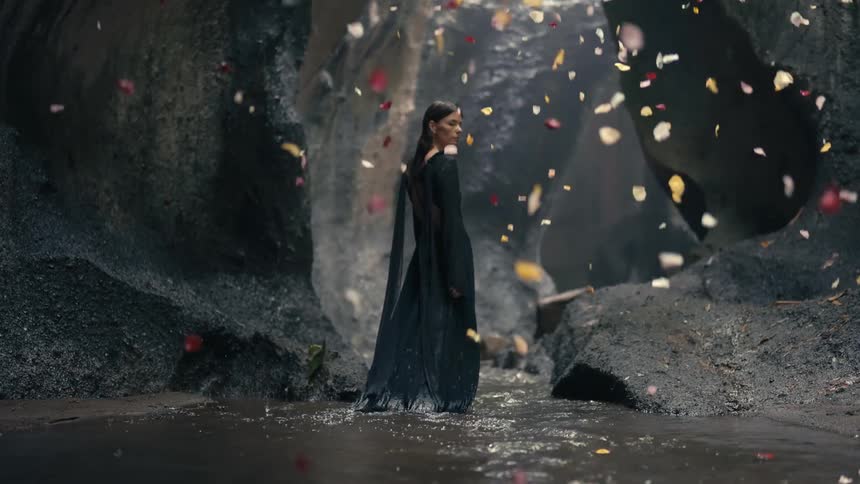 A woman in a long gown is walking through water in a canyon or cave structure as petals fall from above