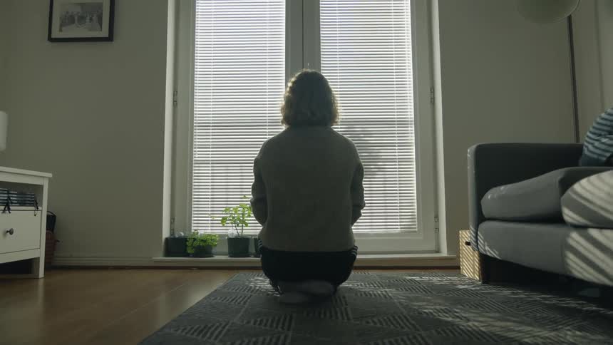 A woman sits on the floor of her living room and looks out the window